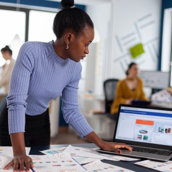 African woman scorlling on laptop looking concentrated at charts and multiethnic colleagues working on marketing. Diverse team of business people analyzing company financial reports from computer.